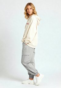 Felpa beige con testo ricamato rosso, abbinata a pantaloni cargo grigi con tasche laterali e polsini elasticizzati, completata da sneakers bianche.