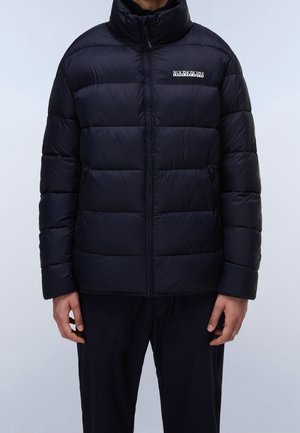 Mann trägt eine schwarze Steppjacke mit durchgehendem Reißverschluss und sichtbarem Markenlogo auf der Brust sowie schwarze Hosen, steht vor einem schlichten Hintergrund.