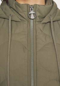 Oliwkowa pikowana kurtka z wysokim kołnierzem, szarym zamkiem błyskawicznym i regulowanym kapturem. Na tkaninie widoczne subtelne wzory faliste.