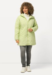 Chaqueta verde acolchada con patrones de hojas, cierre frontal, cuello alto y bolsillos, llevada sobre un suéter de cuello alto blanco y jeans grises. Zapatillas completan el look.