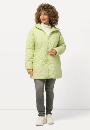 Quilted green jacket with leaf patterns, zip front, high collar, and pockets, worn over a white turtleneck and gray jeans. Sneakers complete the look.