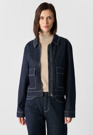 Dark denim cropped jacket with a collar, two front pockets, and contrasting white stitching, worn over a beige turtleneck sweater.