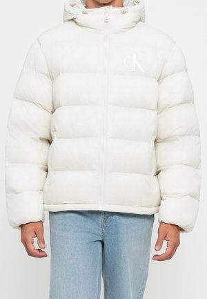 Veste matelassée blanche avec capuche, fermeture éclair à l'avant et logo CK, portée par une personne en jean bleu clair, debout devant un fond uni.