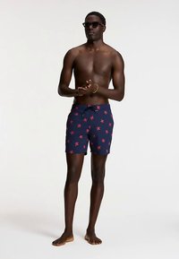 Pantaloni scurți de înot bleumarin cu model de stea de mare roșie, talie elastică cu șnur și croială lejeră. Modelul stă desculț, purtând ochelari de soare.