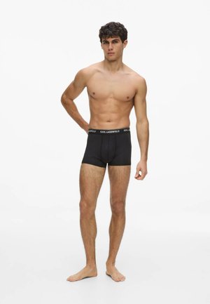 Männliches Model steht barfuß in schwarzen Karl Lagerfeld Boxershorts und posiert mit einer Hand in der Hüfte vor weißem Hintergrund.