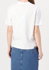 Vit bomulls-T-shirt med korta ärmar och rund halsringning, har en svart logotypetikett vid fållen, kombinerad med en blå jeanskjol.