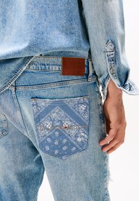 Hellblaue Jeans aus Denim mit einer gemusterten Gesäßtasche, einem braunen Lederlabel mit "TOMMY JEANS" und sichtbaren Steppnähten.