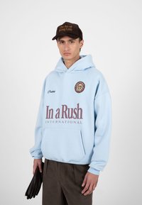 Jeune homme portant un sweat à capuche bleu clair avec le texte « In a Rush International », une casquette marron avec « The Art of Being Late », et tenant des gants noirs.