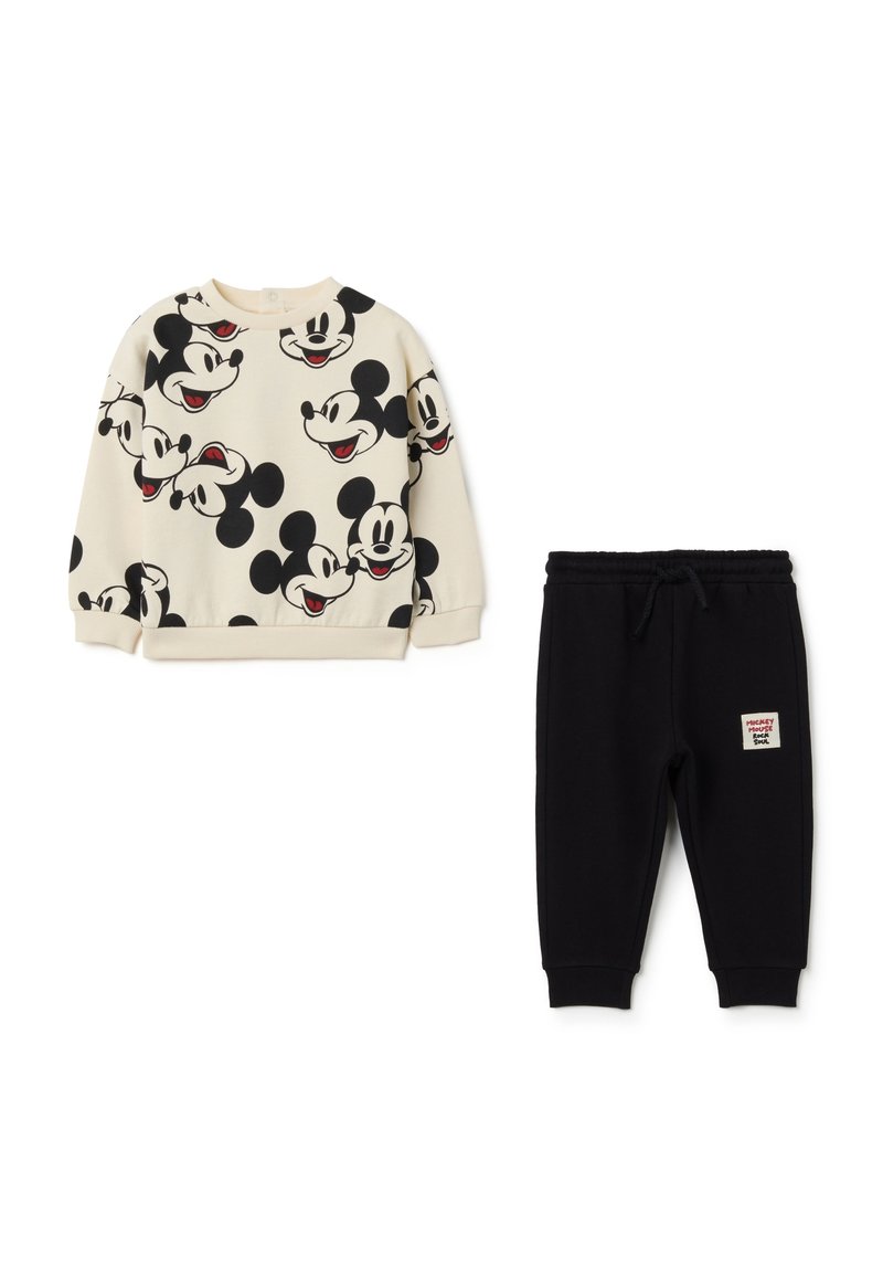 Sweatshirt crème avec des visages de Mickey Mouse noirs, associé à un pantalon de jogging noir avec une étiquette. Manches et taille côtelées, tissu doux.