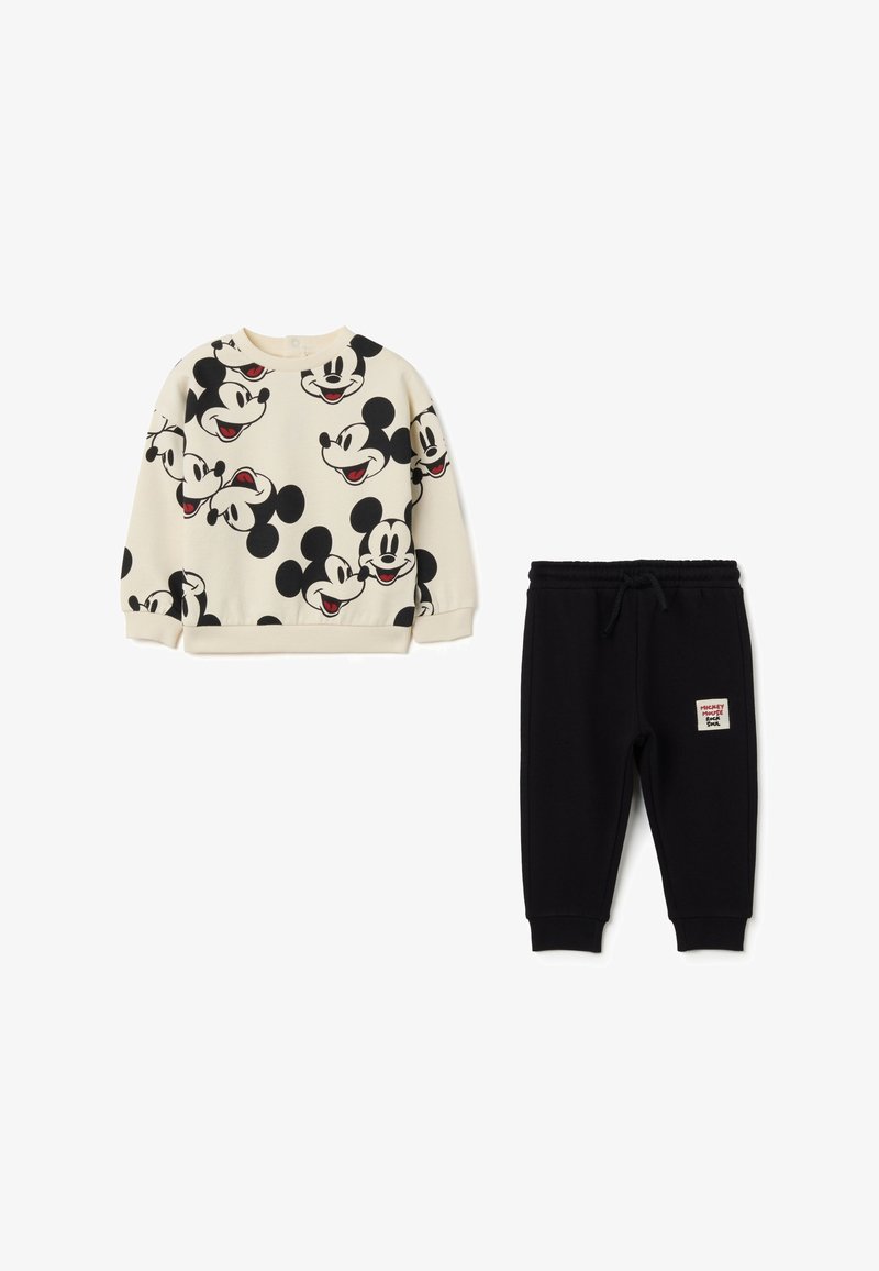 Cremefarbener Sweatshirt mit schwarzen Mickey-Maus-Gesichtern, kombiniert mit schwarzen Sweatpants mit einem Label. Rippbündchen und -bund, weicher Stoff.