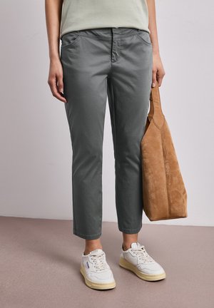 Person iført grå korte bukser, hvide sneakers og holder en brun ruskindstaske mod en ensfarvet baggrund.