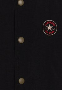 Czarna kurtka o teksturowanym wykończeniu, z brązowymi napami oraz okrągłym, wyszywanym logo "Converse All Star" w kolorze czerwonym i srebrnym.