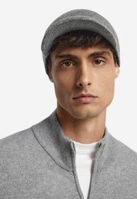 Grå stickad mössa med rundad krona och subtil ribbad textur, bärs tillsammans med en grå zip-up tröja över en vit skjorta.