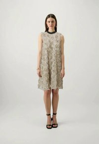 Robe sans manches beige avec une texture florale, arrivant au genou, ornée de roses en tissu superposées. Associée à des talons noirs à lanières et à des bijoux minimalistes.