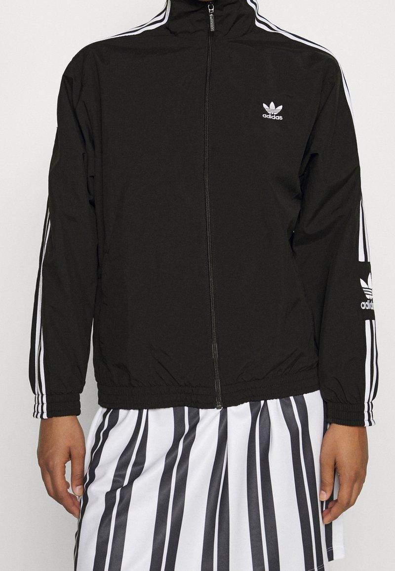 Person som bär en svart Adidas zip-up jacka med vita ränder på ärmarna och en svart-vitt vertikalt randig kjol.