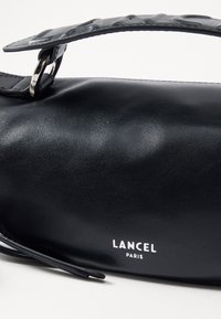 Melnas ādas rokassomas ar teksturētu siksnu, sudraba detaļām, gludu apdari un baltu uzspiesto logo "LANCEL PARIS".