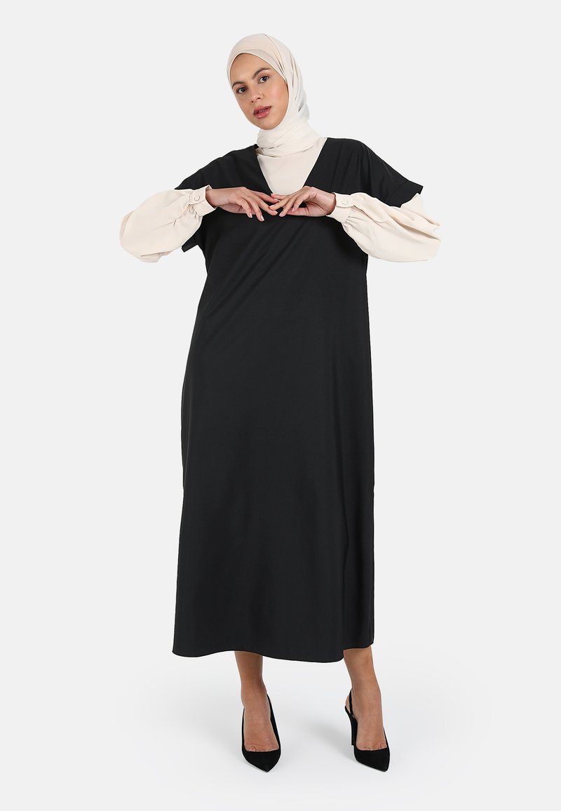 Langes schwarzes Kleid mit kurzen Ärmeln und V-Ausschnitt. Das Model trägt einen beigen Hijab, schwarze Absätze und zeigt eine glatte Stofftextur.