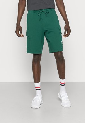 Mann trägt dunkelgrüne kniehohe Cargo-Shorts mit Tunnelzug in der Taille, weiße Socken mit roten und schwarzen Streifen und weiße Turnschuhe.