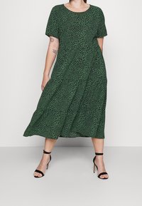 Grünes knielanges Kleid mit kurzen Ärmeln und einem Blättermuster. Hat eine lockere Passform, eine geraffte Taille und eine glatte Textur. Schwarze Absatzsandalen.