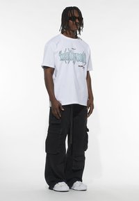 White short-sleeve t-shirt with graphic text, paired with black cargo pants featuring pockets. Model wears dark sunglasses and white sneakers.
