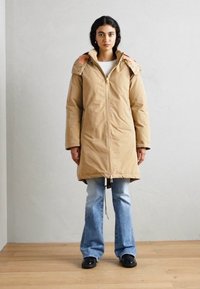 Beige dunjakke med hette, vertikale lommepdekke med glidelås og kontrastfargede kragedetaljer, brukt over lyseblå utsvingte jeans og svarte sko.