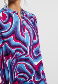 Färgglad blus med ett virvlande mönster i blått, lila och rosa, med ståkrage och delvis dragkedjedetalj.