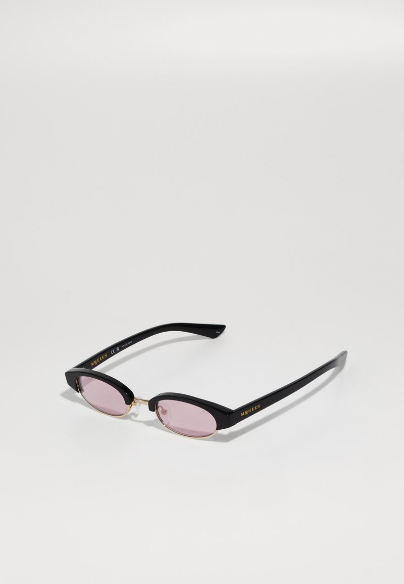 Čierne slnečné okuliare s oválnymi ružovými sklíčkami a zlatými detailmi, logo "MCQUEEN" v zlate na stranách, ležiace na bielej ploche.
