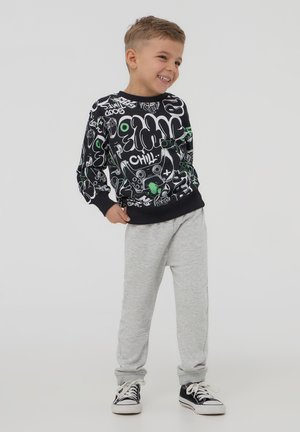 Felpa nera con design grafico bianco e verde, pantaloni della tuta grigi e scarpe nere; presenta polsini ed elastico in vita.