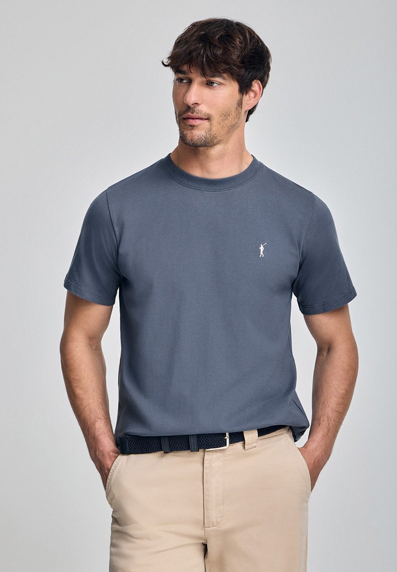 Grijze t-shirt met korte mouwen van zachte katoen, met een ronde halslijn en een klein wit embleem op de borst. G gedragen met lichte beige broek.