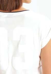 Bílá bavlněná tričko s velkým, mírně vyvýšeným číslem "13" potištěným na zadní straně. Krátké rukávy a kulatý výstřih.