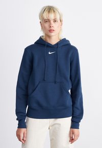 Tamsiai mėlynas Nike pūstinis megztinis su priekinėmis kišenėmis, raišteliu ir gumuotais rankogaliais. Ant krūtinės yra baltas logotipas. Minkšta audinio tekstūra.