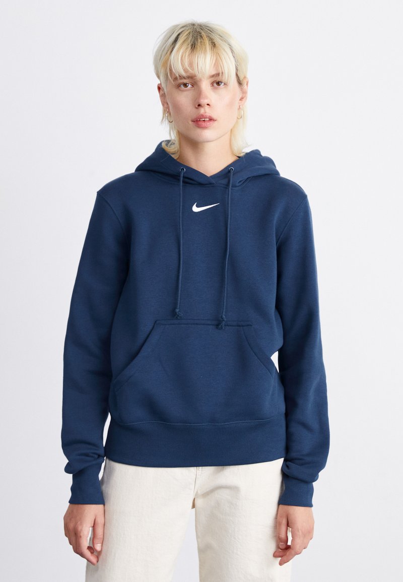 Tamsiai mėlynas Nike pūstinis megztinis su priekinėmis kišenėmis, raišteliu ir gumuotais rankogaliais. Ant krūtinės yra baltas logotipas. Minkšta audinio tekstūra.