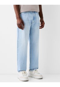 Man som bär ljusblå rakbenade jeans och vita sneakers, står mot en enkel vit bakgrund.