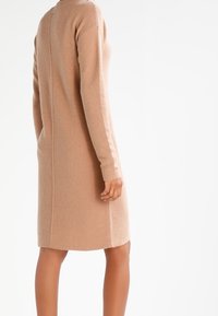 En modell som bär en lång, beige stickad klänning står med ryggen mot kameran och visar upp klänningens texturerade tyg och sömdetaljer.