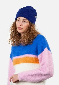 Blue knitted beanie with a textured finish. Model wears a multicolored fuzzy sweater featuring blue, orange, pink, and white stripes.
