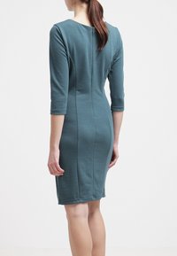 Robe ajustée longueur genou en tissu turquoise, avec des manches trois-quarts, un col rond et un détail de couture courant verticalement dans le dos.