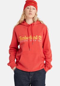 Sudadera de algodón roja con un bolsillo tipo canguro, con "Timberland" y el logo impresos en amarillo. Combinada con un gorro rojo a juego.