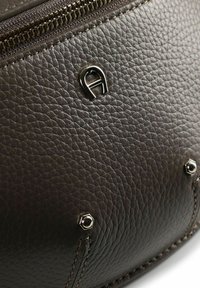 Bolso de cuero marrón oscuro con textura que cuenta con cierre de cremallera y logo en relieve. Incluye herrajes metálicos brillantes y detalles de costuras reforzadas.