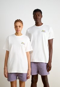 Weiße Rundhals-T-Shirts mit buntem "On Vacation"-Stickmuster. Modelle tragen pastelllila Shorts mit strukturiertem Muster.