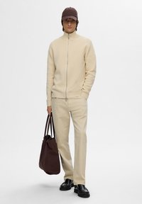 Helle beige Zip-up-Strickjacke mit gerippter Textur, beige Hose, schwarze Loafers und eine braune Mütze; hält eine dunkelbraune Tragetasche.