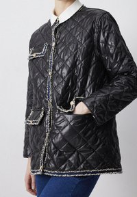 Kvindelig iført en sort quiltet jakke med hvid krave, dekorativ kant og guldknapper, parret med blå jeans.