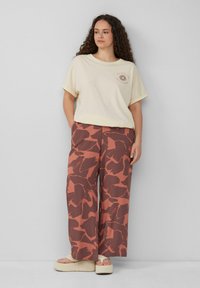 Cremfarbener, übergroßer T-Shirt mit umgeschlagenen Ärmeln und Sonnenmotiv; weite Hosen in Rostfarbe mit abstraktem Muster; Plateausandalen.