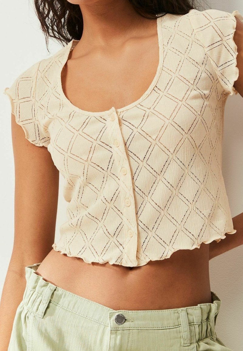 Femme portant un haut beige à manches courtes boutonné avec un motif géométrique en losange et un pantalon taille haute vert clair.