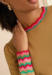 Lichtbruine top met ribbelig, meerkleurig golftrim in rood, groen, roze en beige op de kraag en mouwen; textuurdetail van de stof.
