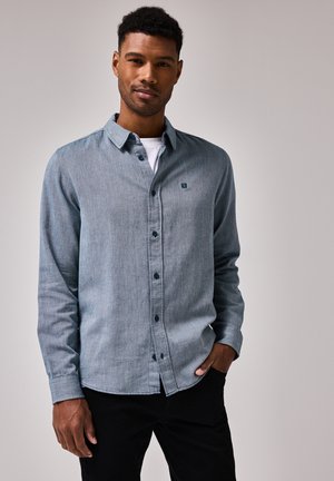 Chemise bleue à manches longues avec boutons, fabriquée à partir d'un tissu texturé, dotée d'un petit patch logo et d'un col classique, associée à un pantalon noir.