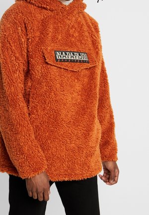 Person wearing an orange fuzzy hooded sweatshirt with a front pocket labeled "Napapijri geographic," paired with black pants.