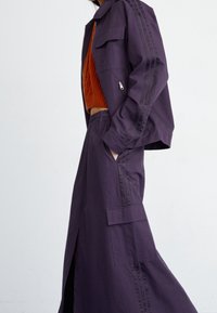 Violetti takki, jossa on vetoketju ja kaksi sivutaskua, yhdistettynä teksturoituun oranssiin lyhytvillatakkiin ja sopivan violettiin hameeseen, jossa on halkioita.