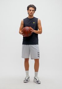 Νεαρός άνδρας με μαύρο αμάνικο μπλουζάκι και γκρι σορτς κρατάει μια μπάλα μπάσκετ, στέκεται σε λευκό φόντο με λευκά αθλητικά παπούτσια.