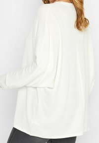 Bílé oversized svetr s žebrovanou strukturou, širokými rukávy a bočními rozparky, spárovaný s tmavě šedými kalhotami. Jednoduchý a volný střih.