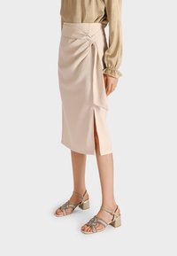 Gonna beige a lunghezza ginocchio con design a portafoglio, caratterizzata da uno spacco laterale e una texture liscia. Abbinata a sandali con tacco a blocco metallici.